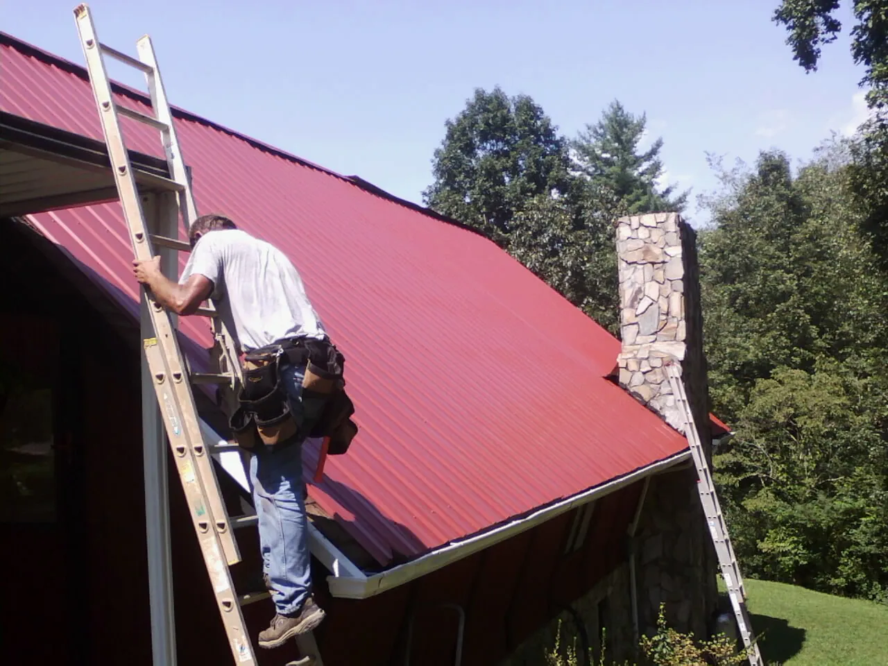Roofer on a ladder beside a red metal roof and stone chimney — Soffit & Fascia Repair by Roofing Brothers in Speed